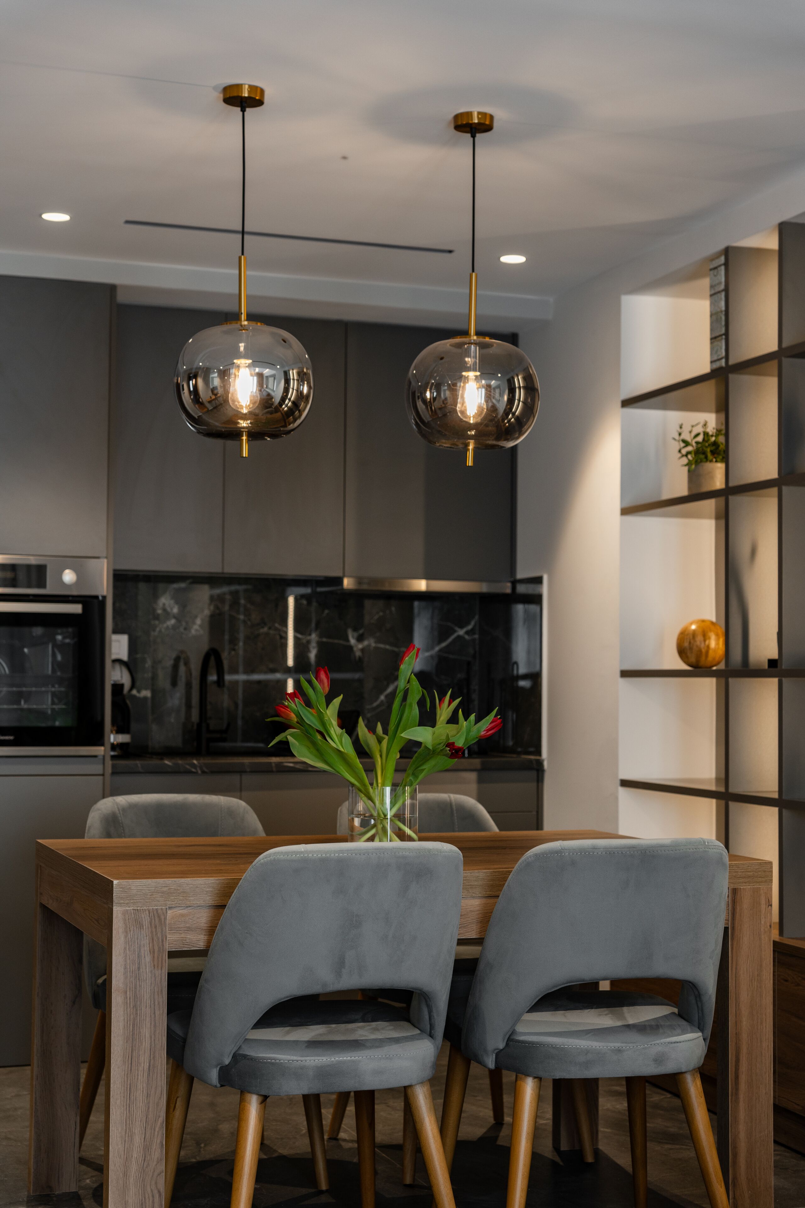 A modern dining area with a wooden table and two grey upholstered chairs. Two pendant lights hang above the table, and a vase with red flowers is on the table.