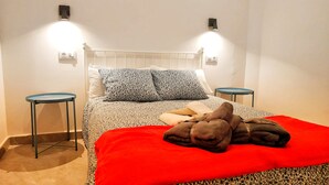 Una cama con una manta roja, dos almohadas estampadas y una toalla marrón. Hay dos mesitas de noche junto a la cama, cada una con una lámpara.