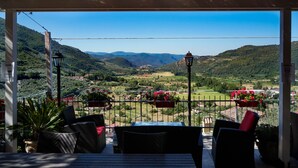 A covered outdoor seating area with wicker furniture and potted plants, overlooking a valley with mountains and a clear sky. - Perla House (Ferentillo)