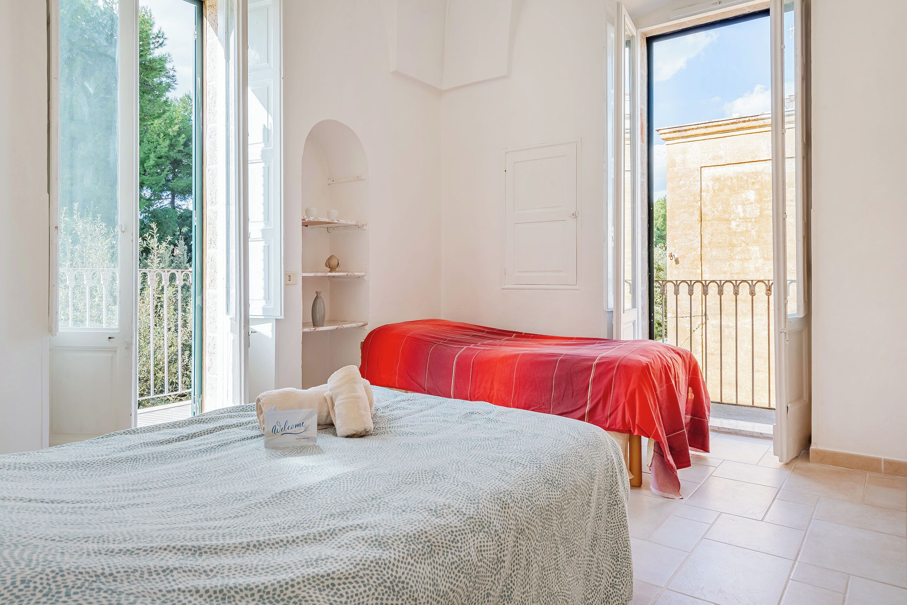 A bedroom with a bed, a red bedspread, a white wall, a door, and a balcony with a railing.
