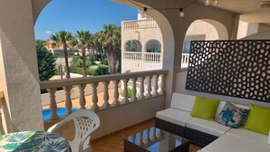 Ein Balkon mit einer weißen Couch, einem Glastisch und einem weißen Stuhl. Im Hintergrund ist ein Pool und Palmen zu sehen.