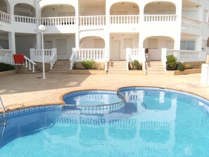 A swimming pool with a sunken center, surrounded by a white building with balconies and a covered staircase.