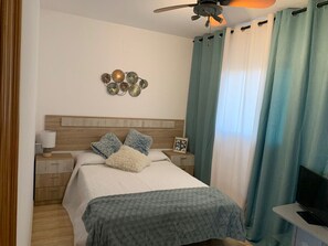 A bedroom with a bed, a nightstand, a wall decoration, a ceiling fan, and curtains.