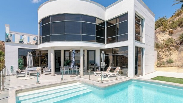 Un bâtiment moderne à deux étages doté d’une piscine, avec une façade en verre incurvé et un espace extérieur aménagé pour s’asseoir.