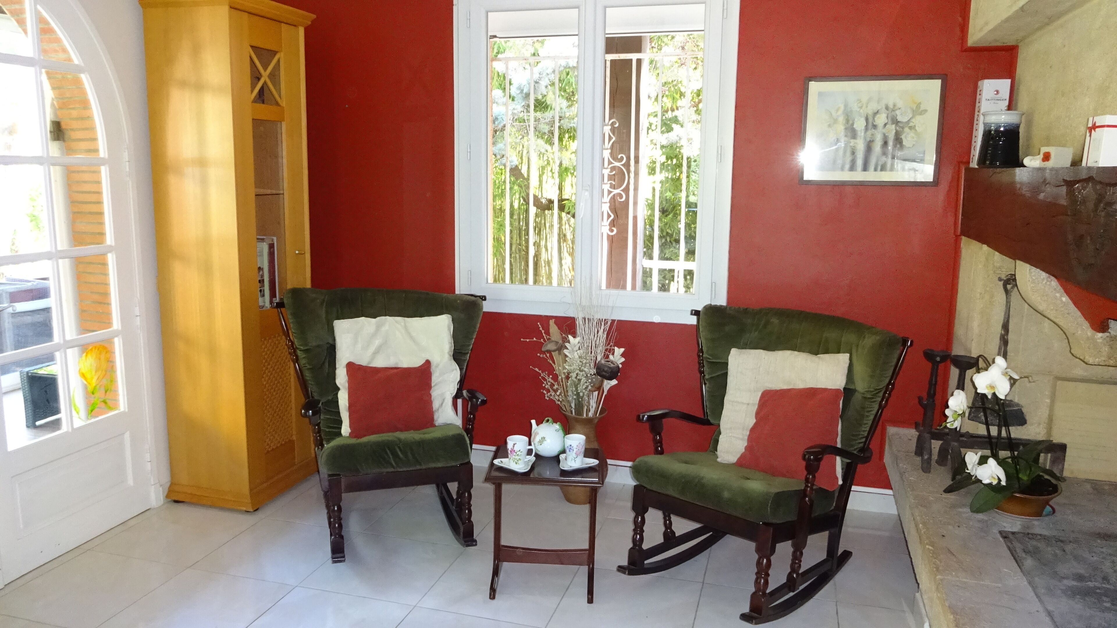 Una habitación con dos sillones tapizados en verde, una mesita con tazas de té, un ventanal de marco blanco y una pared roja.