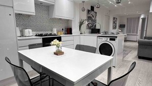 A modern kitchen with white cabinets, a dining table with chairs, a washing machine, and a grey sofa in the living area.