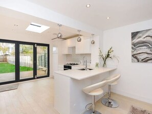 A modern kitchen with a central island, bar stools, pendant lighting, and a view of a backyard through glass doors. - Luxury 3 Bedroom House in Bromley, South East London (Bromley)
