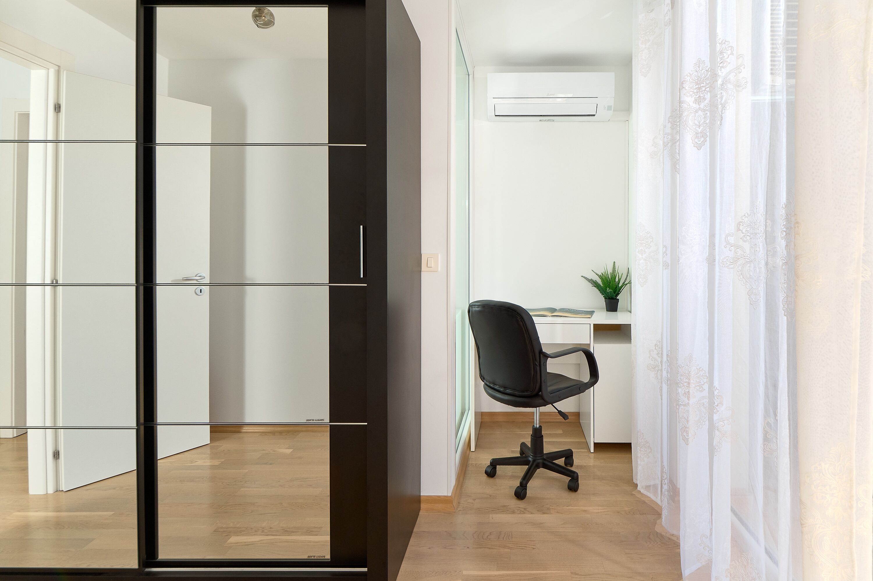 A modern office space with a glass partition, a black office chair, a desk with a computer, and a potted plant.