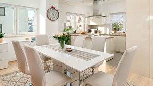 A modern kitchen with white cabinets, a microwave, and a sink. There is a dining area with a white table and chairs, and a clock on the wall. - Sun Spalato Flower (Split)