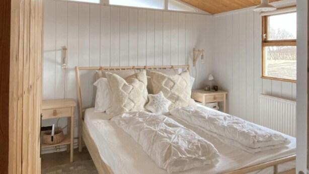 Un dormitorio con cama de madera, paredes blancas, techo inclinado, una mesita de noche y una ventana.