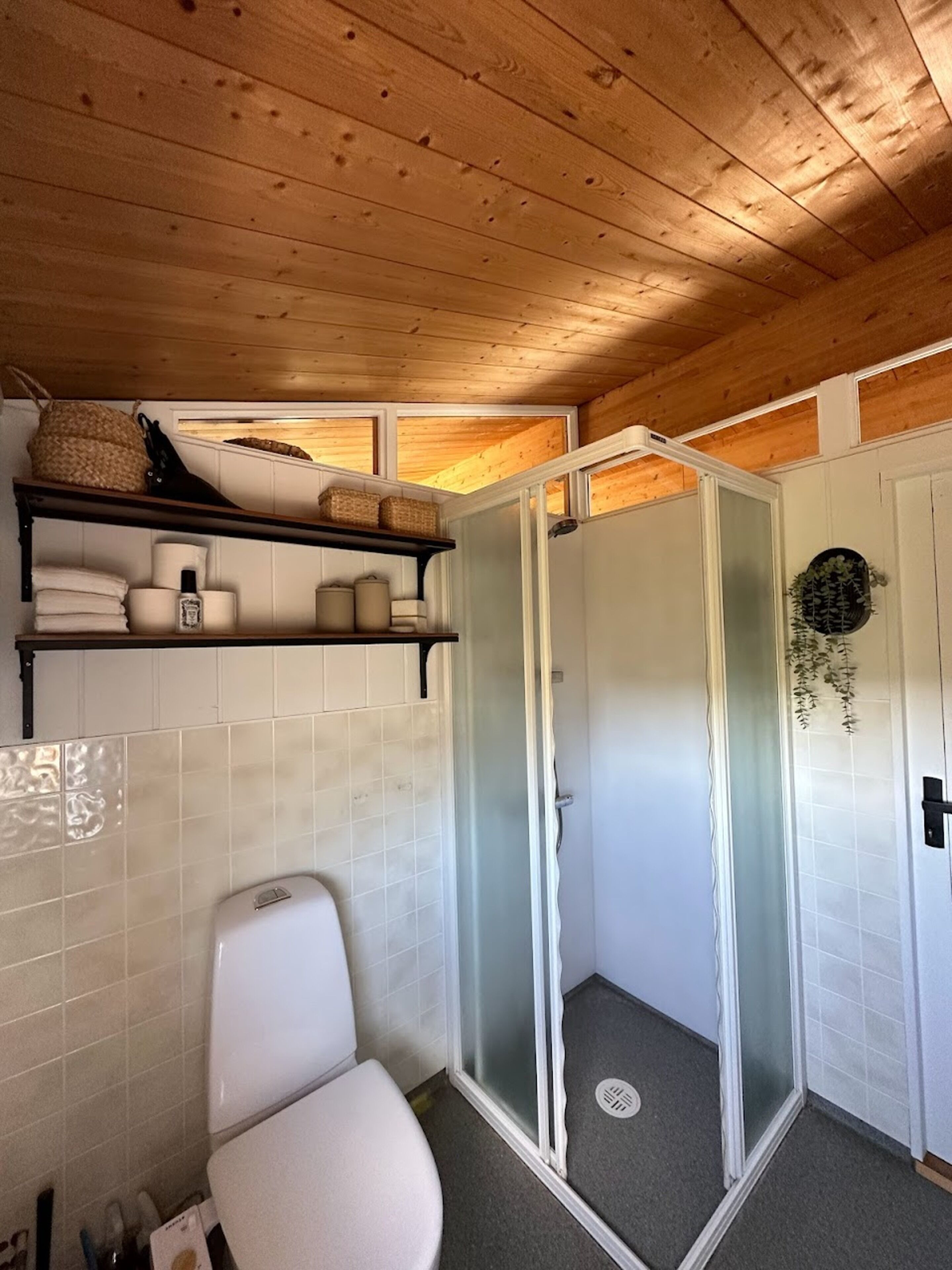A wooden-paneled bathroom with a toilet, a shower enclosure, and a shelf with toiletries.