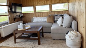 Interior acogedor de una cabaña con sofá, una mesa de centro, televisor montado en la pared y ventana con vista a un paisaje cubierto de hierba.
