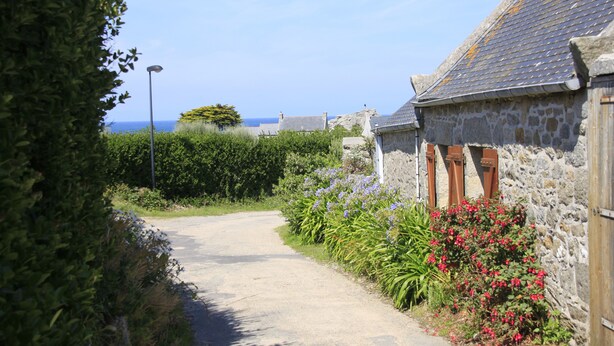 A narrow stone pathway leading to a stone building with a tiled roof, surrounded by lush greenery and flowering plants. - Appartement Neuf, à 5minutes de la Plage, Commerce au Rdc, Pour 4 Personnes (Plounéour-Brignogan-plages)