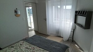 A bedroom with a bed, a bedside table, a mirror, a window with curtains, and a door leading to another room. - Appartement Neuf, à 5minutes de la Plage, Commerce au Rdc, Pour 4 Personnes (Plounéour-Brignogan-plages)