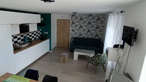 A compact living space with a kitchenette, a sofa, a TV mounted on the wall, and a patterned wallpaper. - Appartement Neuf, à 5minutes de la Plage, Commerce au Rdc, Pour 4 Personnes (Plounéour-Brignogan-plages)
