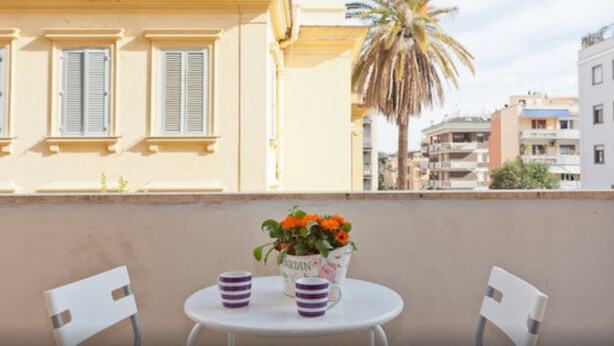 A balcony with a white table and two chairs, a potted plant with orange flowers, and a view of buildings and a palm tree. - Carlo's welcome in Trastevere (Roma)