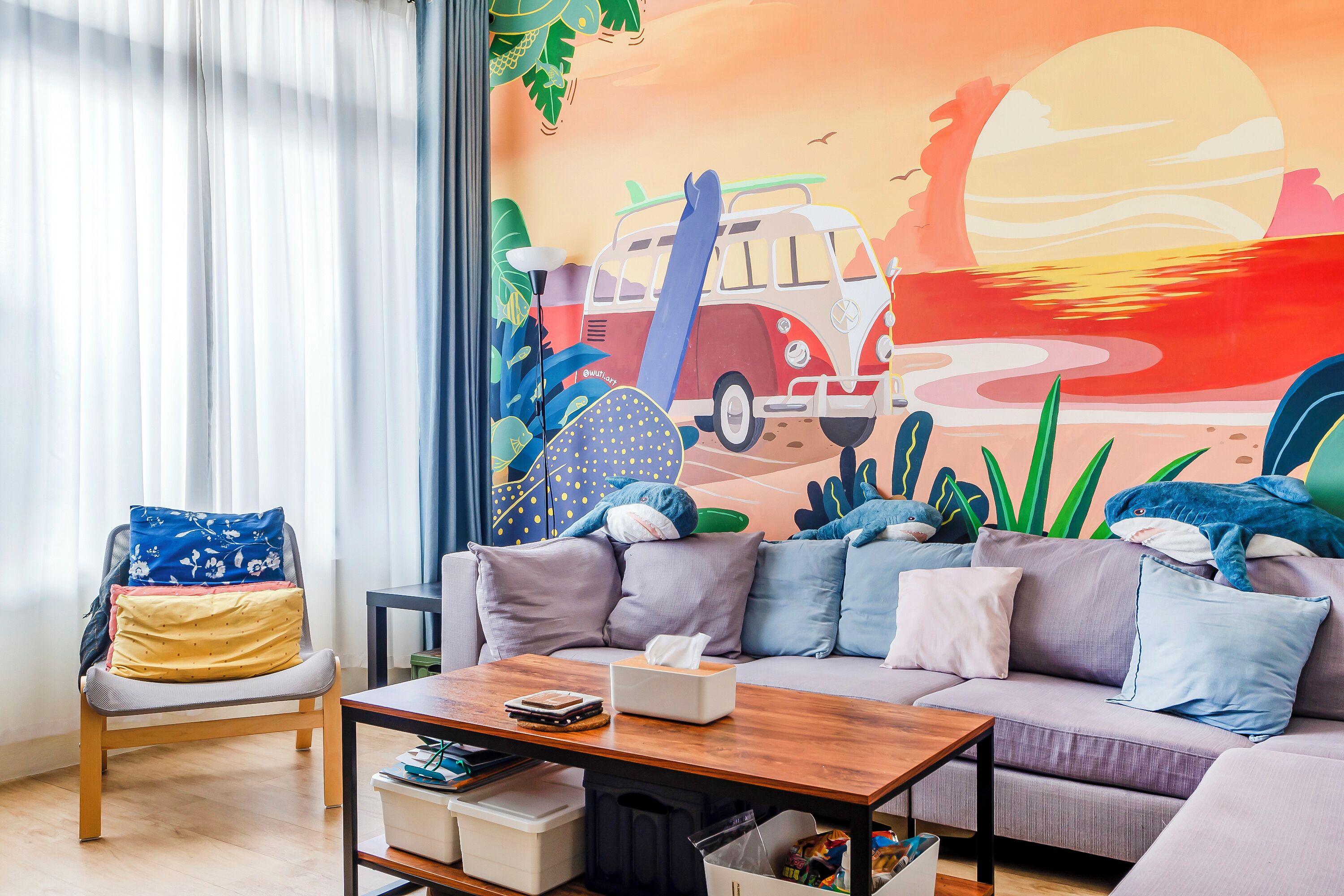 A living room with a sofa, a coffee table, and a chair. There is a mural on the wall depicting a van, surfboard, and tropical plants.