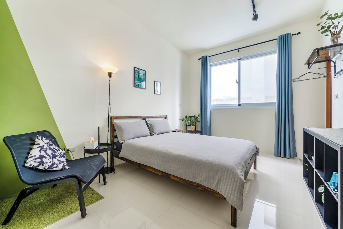 A bedroom with a bed, a chair, a bedside table, a lamp, a window with curtains, and a plant.