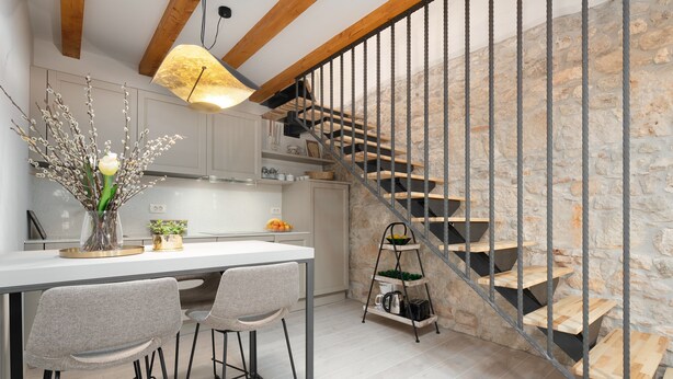 Una cocina moderna con encimera blanca, dos taburetes de bar, un florero con ramas y una escalera con paredes de piedra y barandillas de metal.