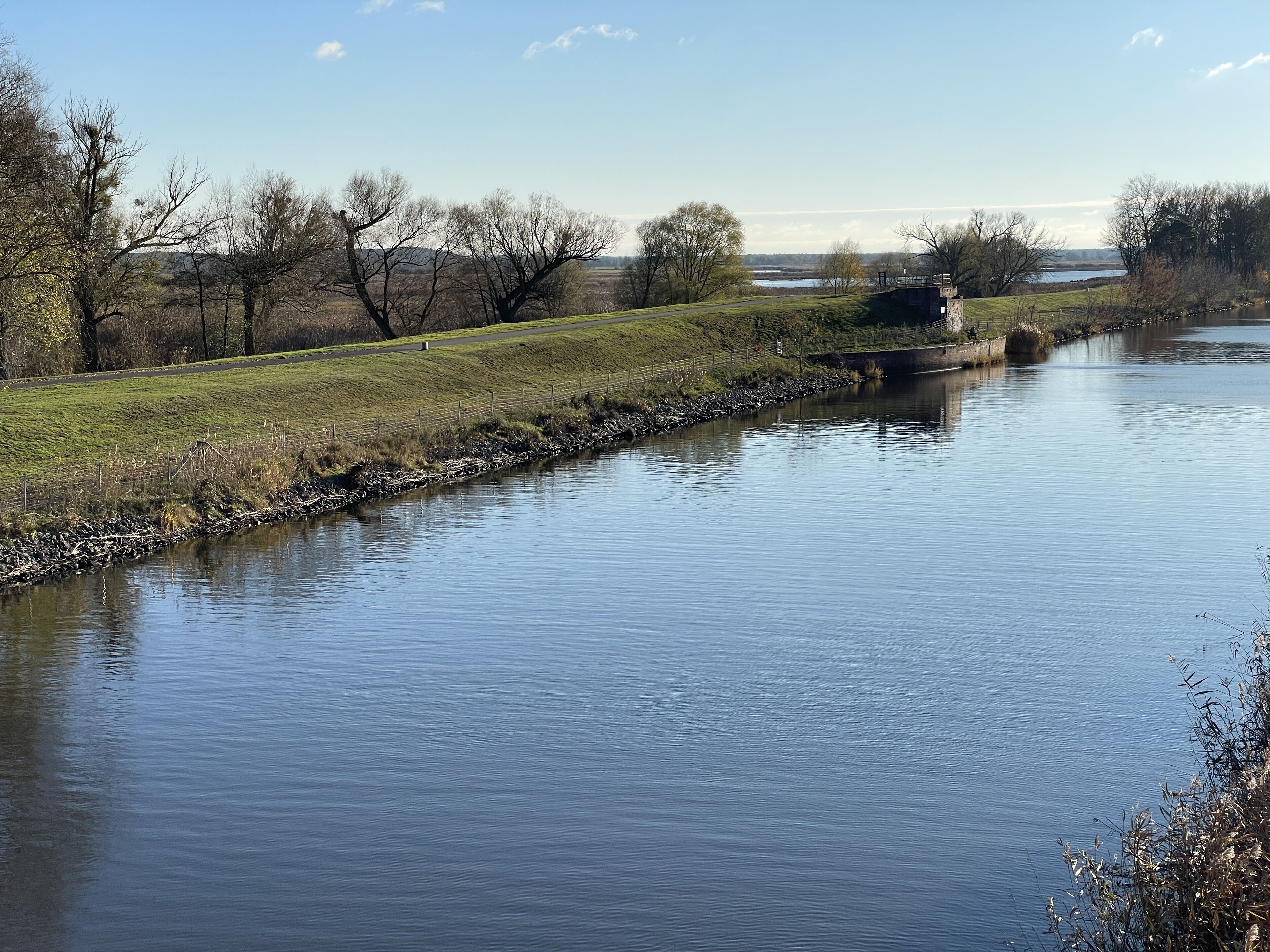 A calm river with a stone embankment, grassy banks, and trees in the background.