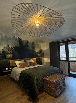 A bedroom with a large bed, a headboard with a forest mural, a hanging light fixture, and a large window. - - La Tanière - (La Broque)