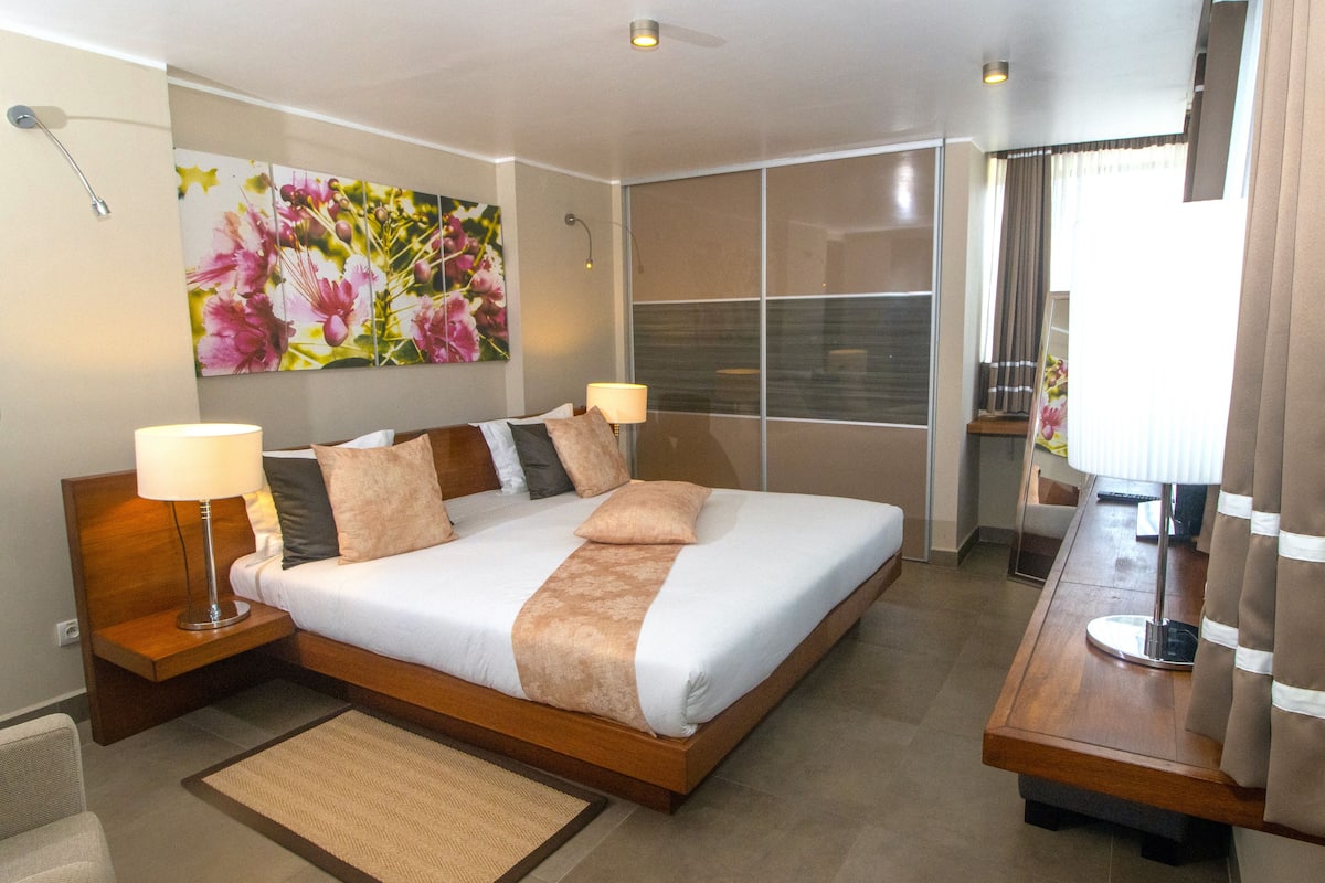 Ein modernes Hotelzimmer mit einem großen Bett, zwei Nachttischlampen, einem Holznachttisch, einem Spiegel und einem großen floralen Kunstwerk an der Wand.