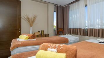 Two beds with neatly folded towels, a small table with a vase of dried plants, and a view through sheer curtains to the outdoors.