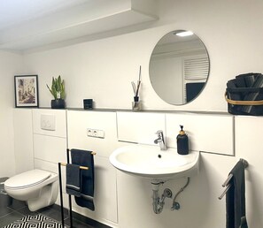 Een moderne badkamer met toilet, wastafel en spiegel. Handdoeken hangen aan een rek en op het aanrecht staat een plant in pot.