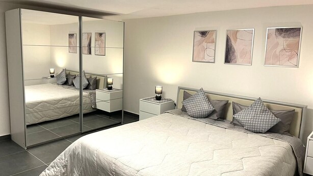A bedroom with a large mirror, a bed with a quilted cover, bedside tables with lamps, and abstract wall art. - Stilvolles Designappaetment mit Terrasse & Garten im Idylischem Bindlach (Bindlach)
