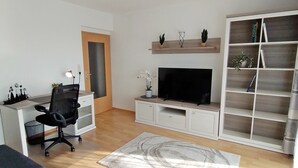 Una sala de estar moderna con un televisor de pantalla plana, una silla de oficina negra, un mueble blanco para televisión, suelo de madera y una amplia estantería.