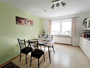 A dining area with a table set for four, a radiator, a window with curtains, and a kitchen area with cabinets and appliances. - 80 qm Moderne Wohnung mit Balkon und Garten im Herzen von Tirschenreuth (Tirschenreuth)