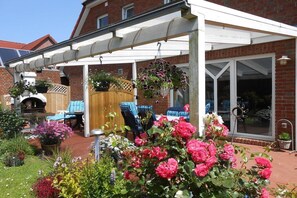 Ein überdachter Patio-Bereich mit Gartenmöbeln, ein Backsteinhaus und ein Garten mit verschiedenen Pflanzen und Blumen.