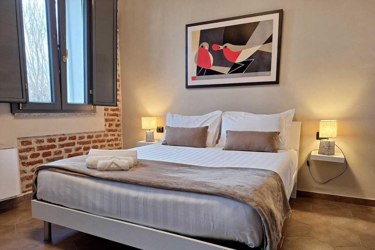 A neatly made bed with a white headboard, two bedside lamps, and a framed abstract painting above it.