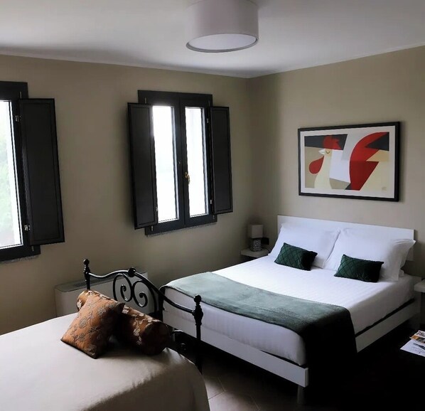 A bedroom with a bed, bedside table, lamp, and a window with shutters. - Cascina Mora Bassa (Vigevano)