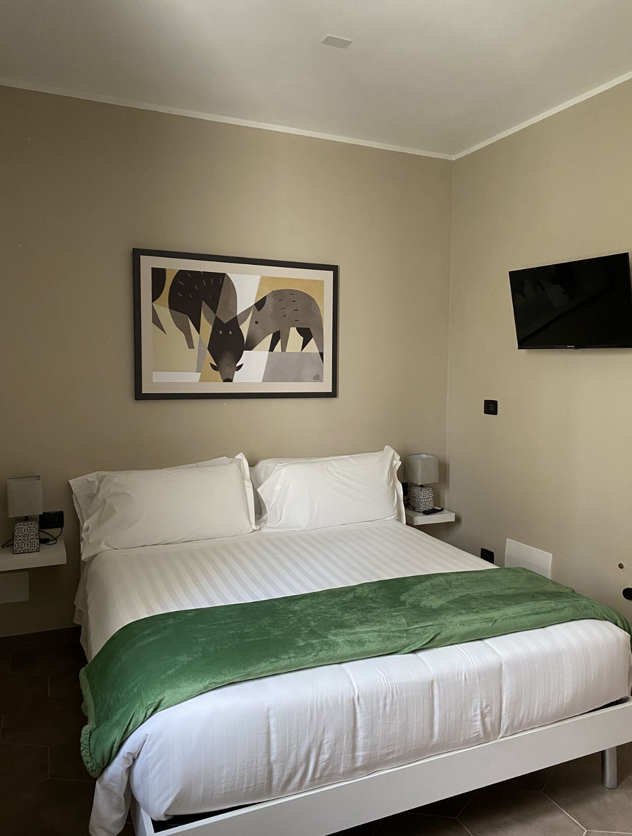Een slaapkamer met een bed, een groene plaid, een lijst met een abstract schilderij, een flatscreen televisie en nachtkastjes met lampen.