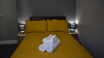 Una cama bien hecha con una colcha amarilla y dos almohadas, un par de toallas dobladas sobre la cama y dos lámparas de noche en mesitas.