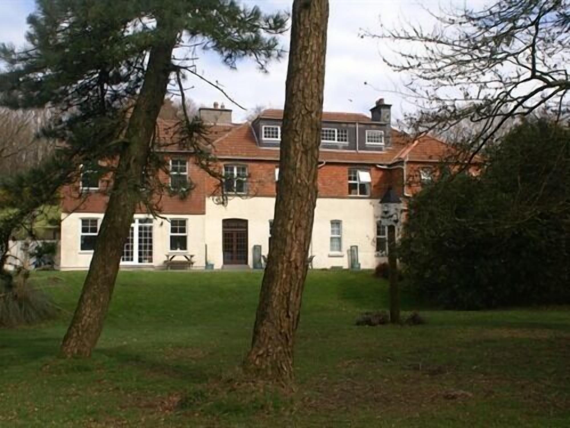 A large, two-story building with a red-tiled roof and white walls, surrounded by trees and a grassy lawn.