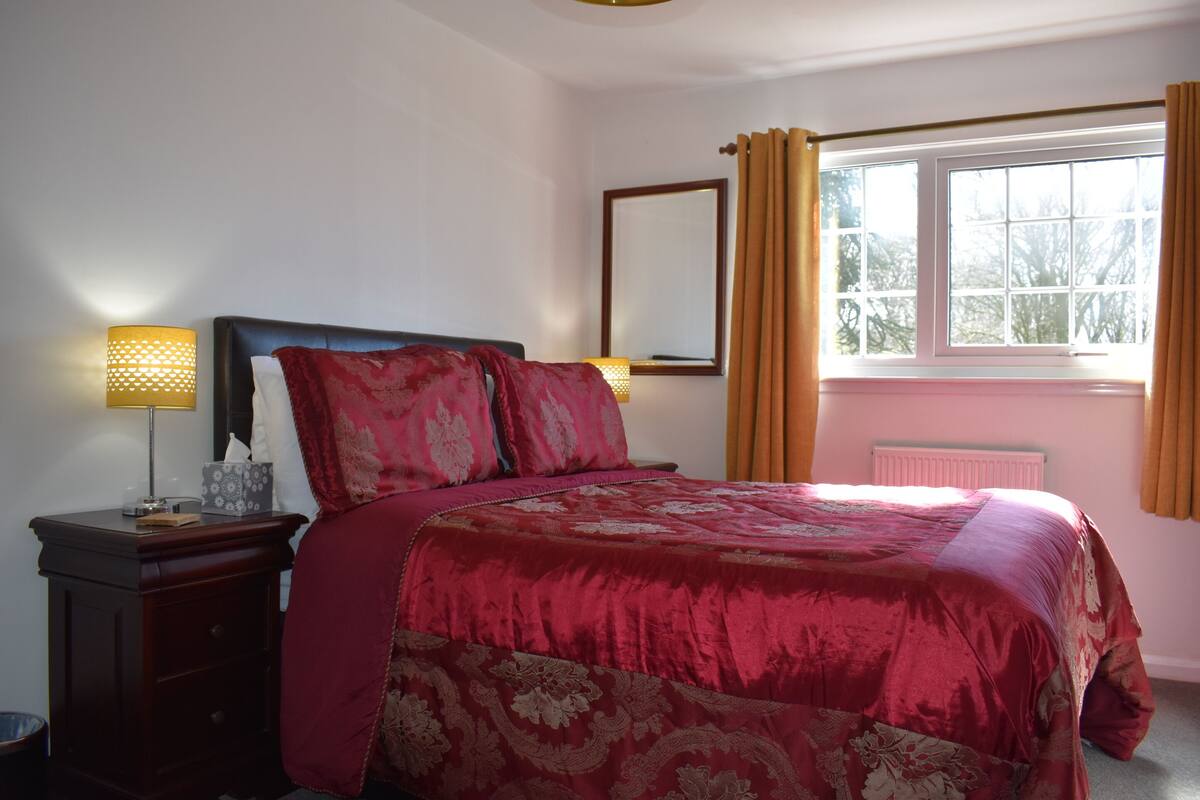 A bedroom with a large bed, a bedside table with a lamp, a window with curtains, and a radiator.