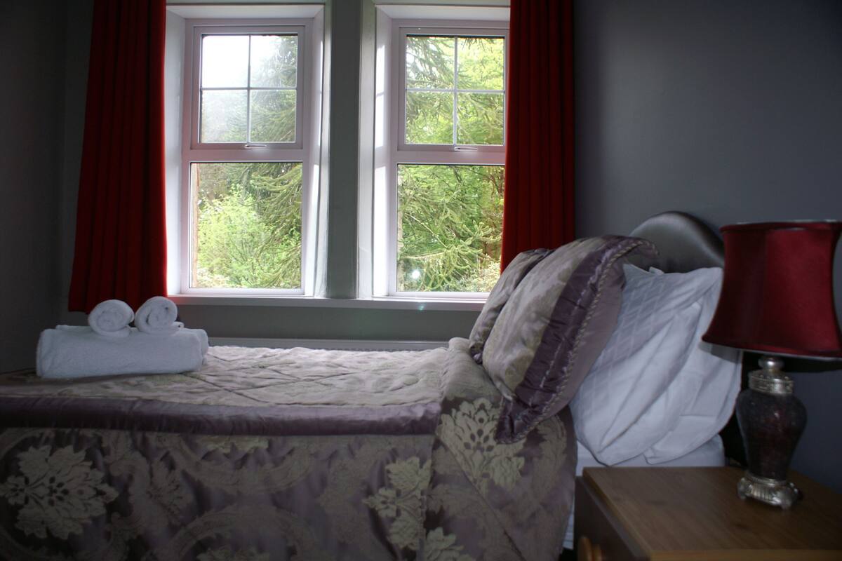 A neatly made bed with a floral bedspread, a bedside table with a lamp, and a window with red curtains.