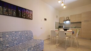 Een moderne woonkamer met een gestreept bankje, een eethoek met stoelen, een keuken met kasten en apparatuur, en een stadsgezicht als kunstwerk aan de muur.