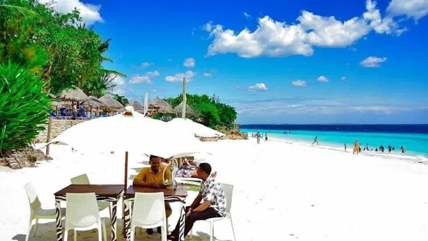A beachside dining area with a table and chairs, a clear view of the ocean, and a tropical setting with palm trees and thatched-roof structures. - COCCOBELLO ZANZIBAR (Kendwa)
