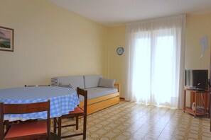 A room with a bed, a table with a blue tablecloth, a TV on a stand, and a window with curtains.