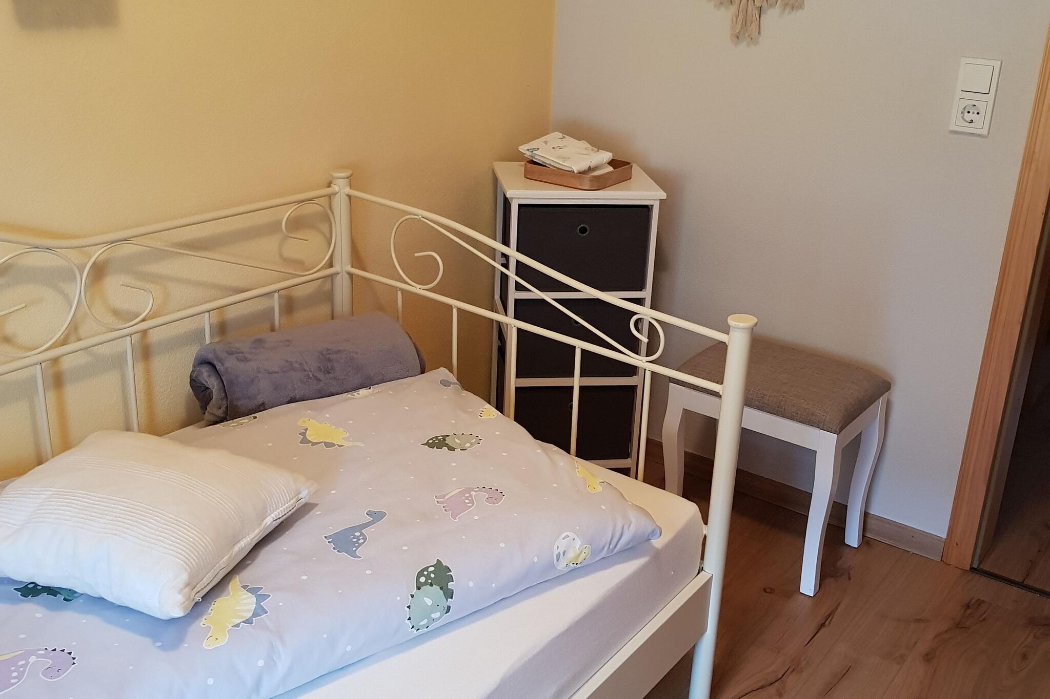 Una cama bien hecha con una colcha estampada y almohadas, una mesita de noche con una lámpara, y un pequeño banco.