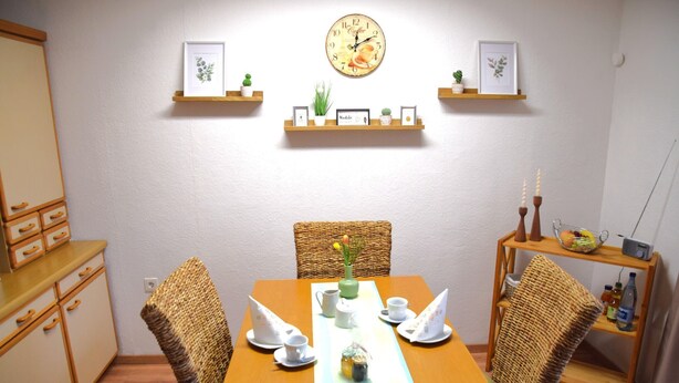 Uma área de refeições com uma mesa de madeira posta para quatro pessoas, rodeada por cadeiras de vime. Existem duas prateleiras fixadas na parede com quadros e pequenas plantas. Um relógio está pendurado na parede acima das prateleiras.