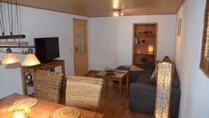 Uma sala de estar aconchegante com teto de madeira, televisão sobre um suporte, cadeiras de vime, um sofá, uma mesa com abajur e uma estante com objetos decorativos.