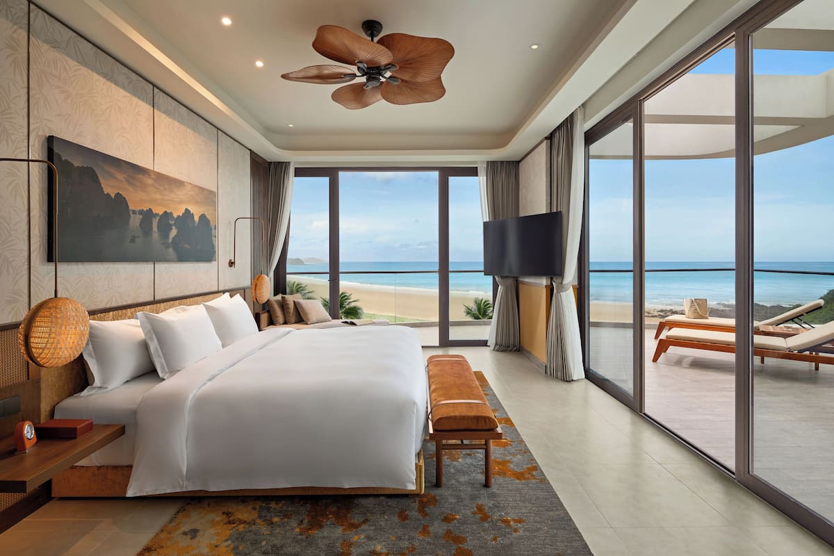 A modern bedroom with a large bed, a ceiling fan, a TV, and a balcony with a view of the beach.