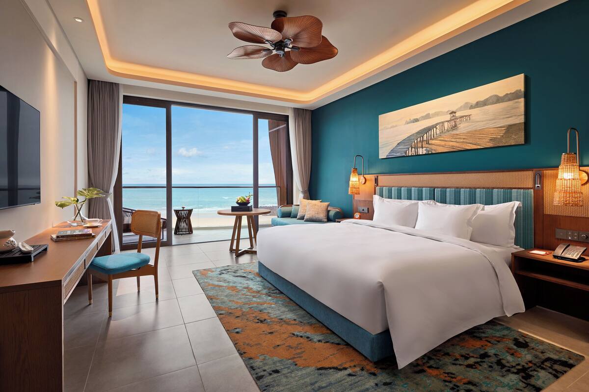 A modern hotel room with a large bed, a desk, and a view of the sea.