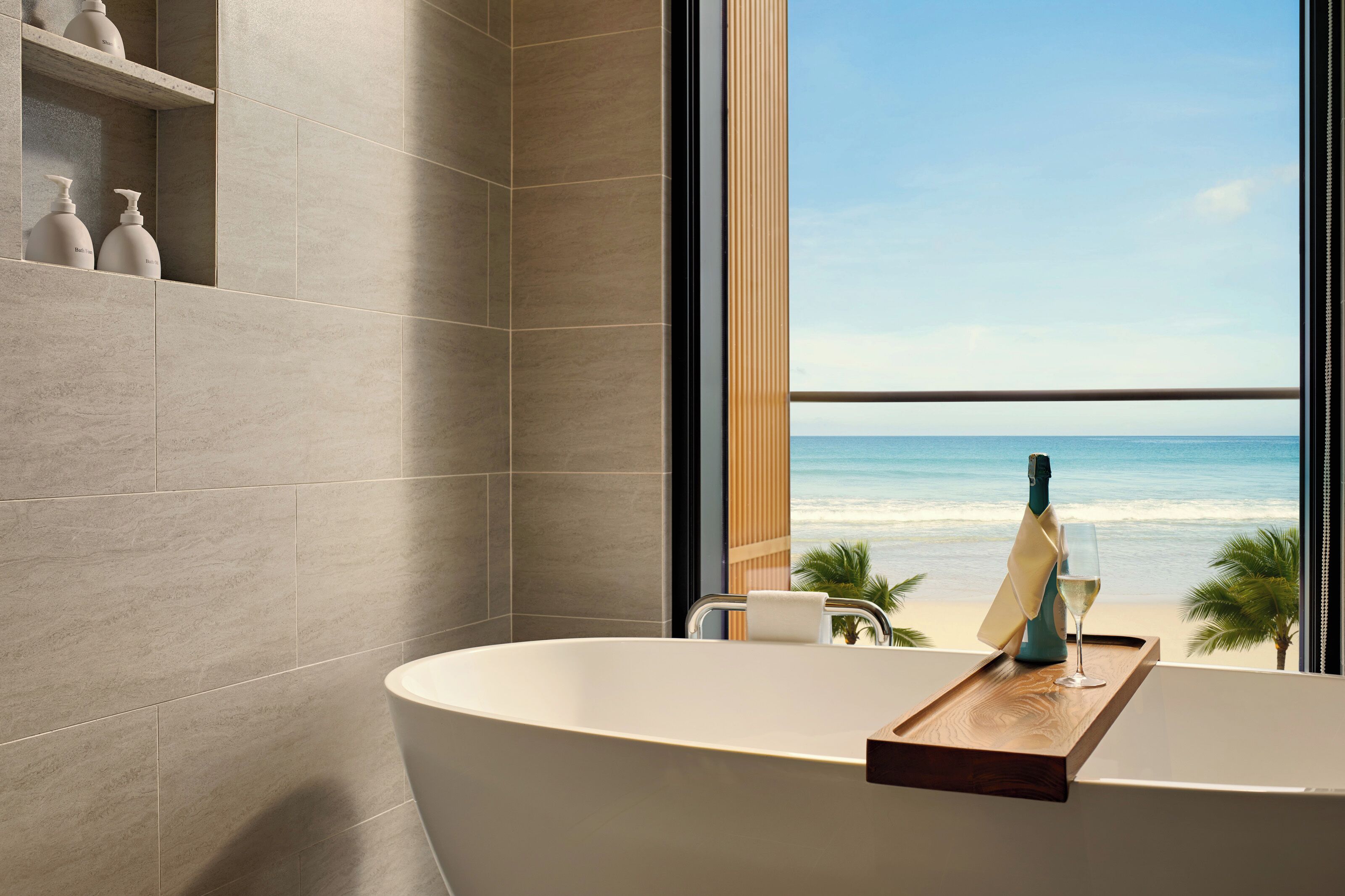 Een moderne badkamer met een vrijstaand bad, een houten dienblad met een fles en glas, en uitzicht op het strand door een groot raam.