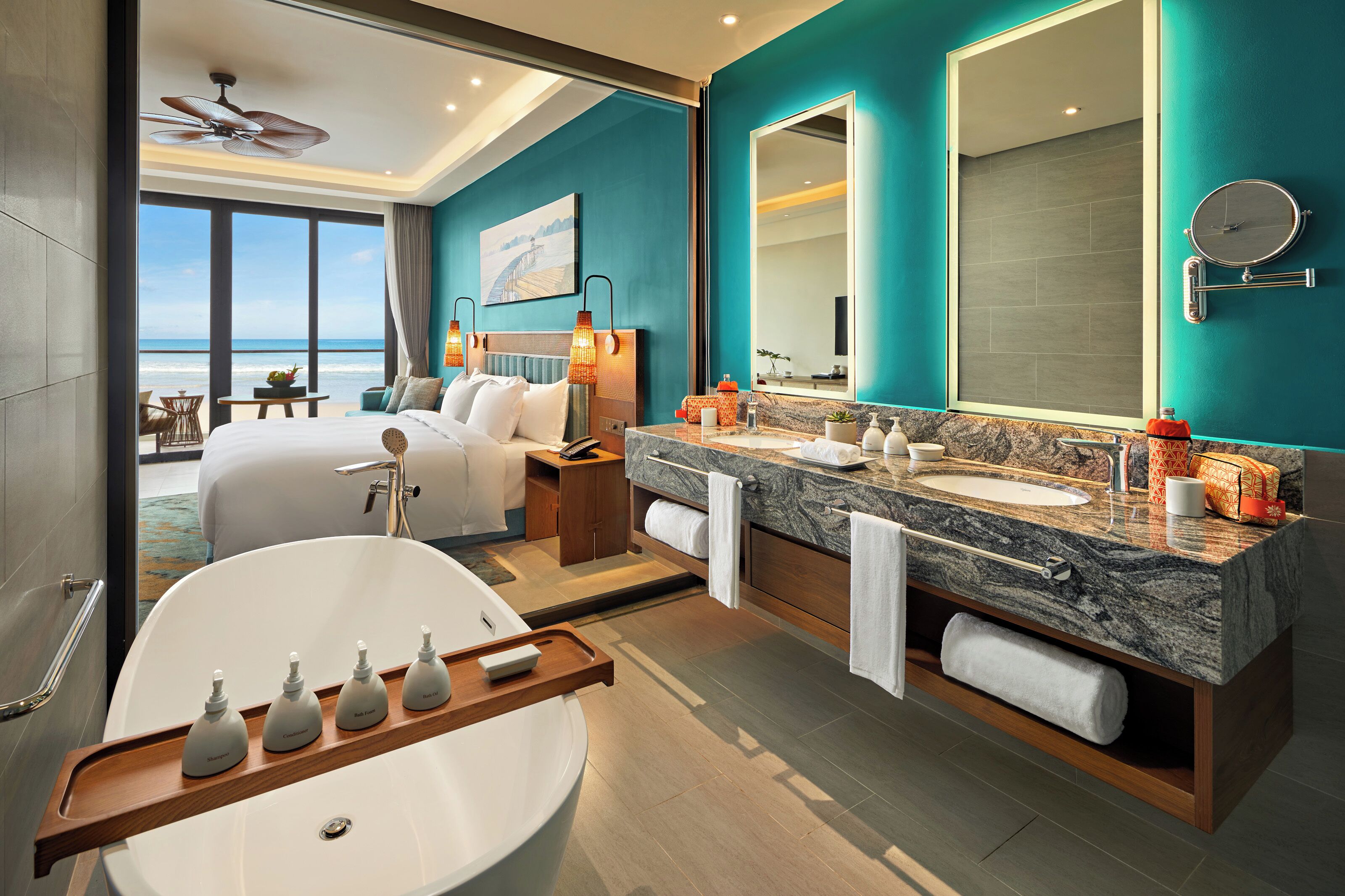 Una habitación de hotel moderna con una cama grande, un baño con bañera y lavamanos doble, y un balcón con vistas al océano.