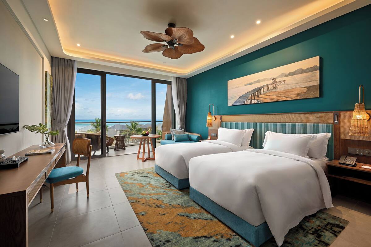 A hotel room with two beds, a desk, a chair, a TV, and a view of the sea.